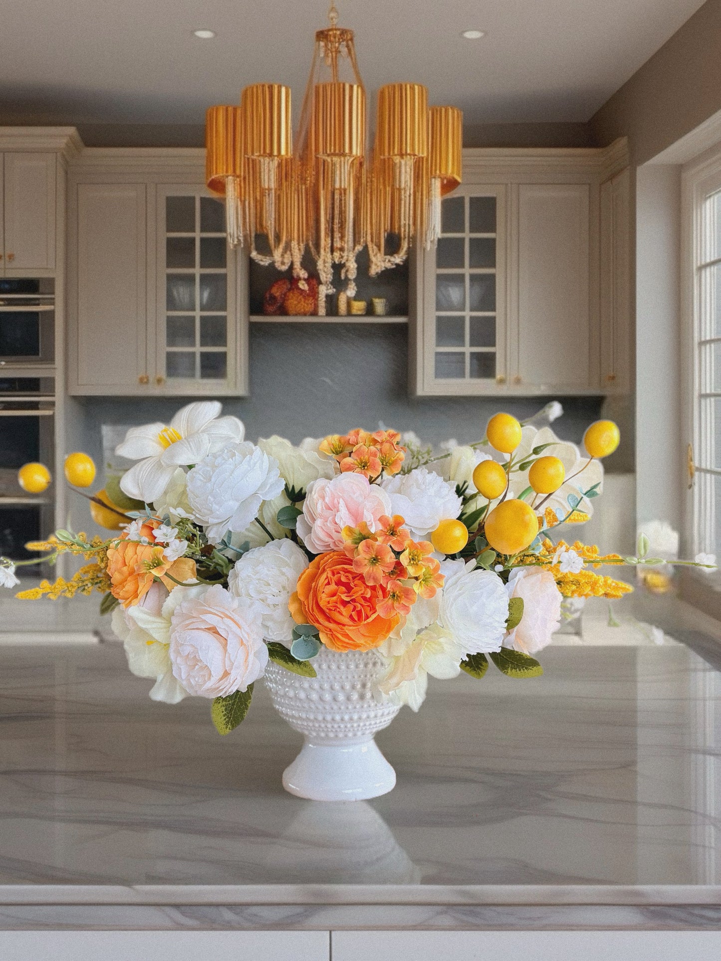 Floral arrangement in a white vase on a kitchen counter with a chandelier above.