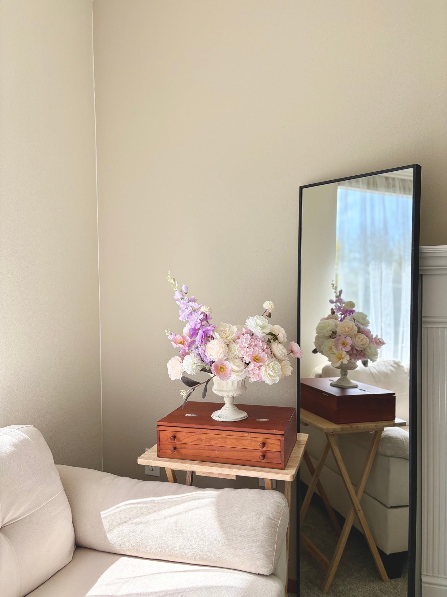 Floral arrangement on a table next to a full-length mirror reflecting a room with a couch.