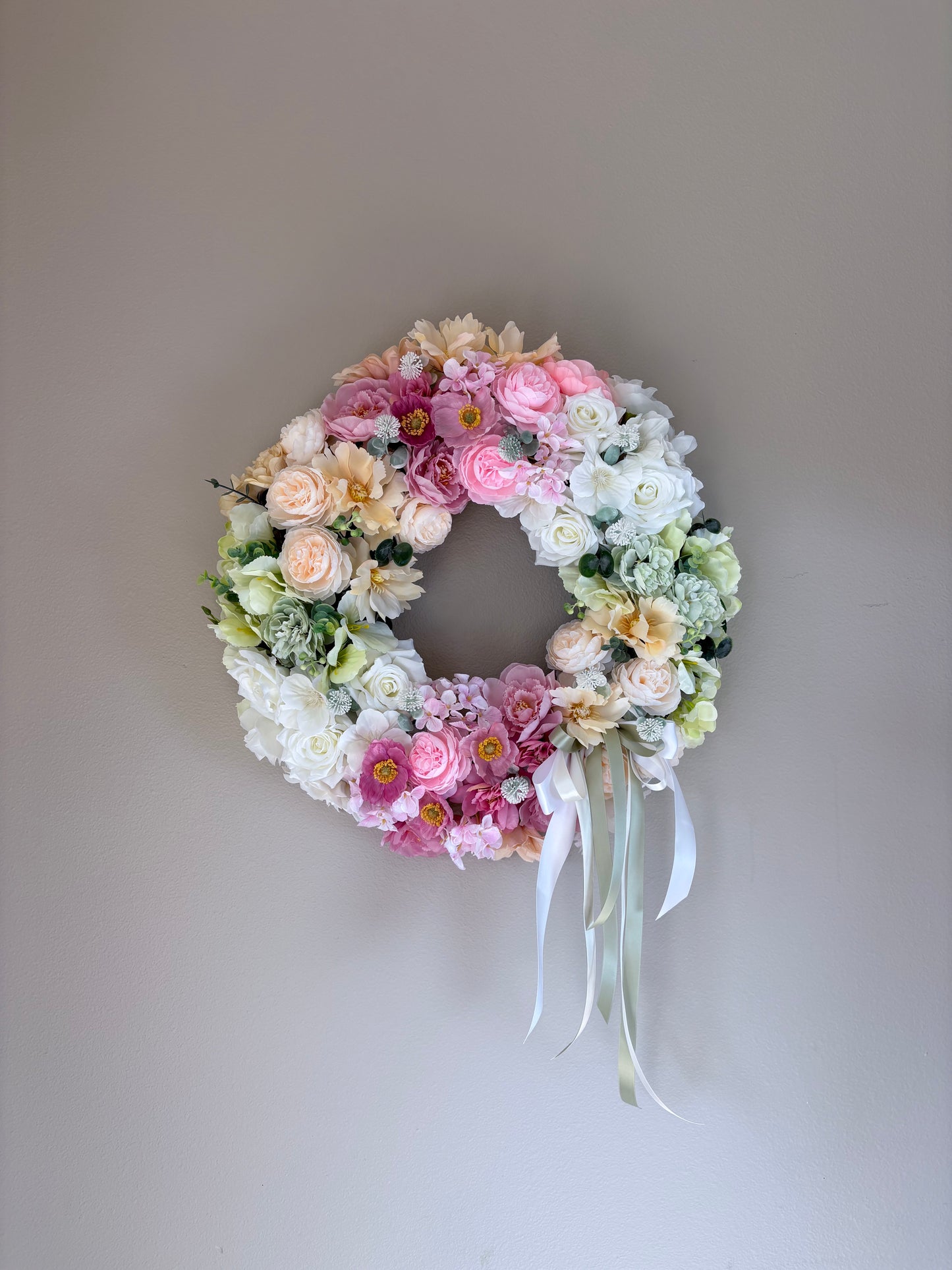 Floral wreath with pink, white, and green flowers on a plain wall.