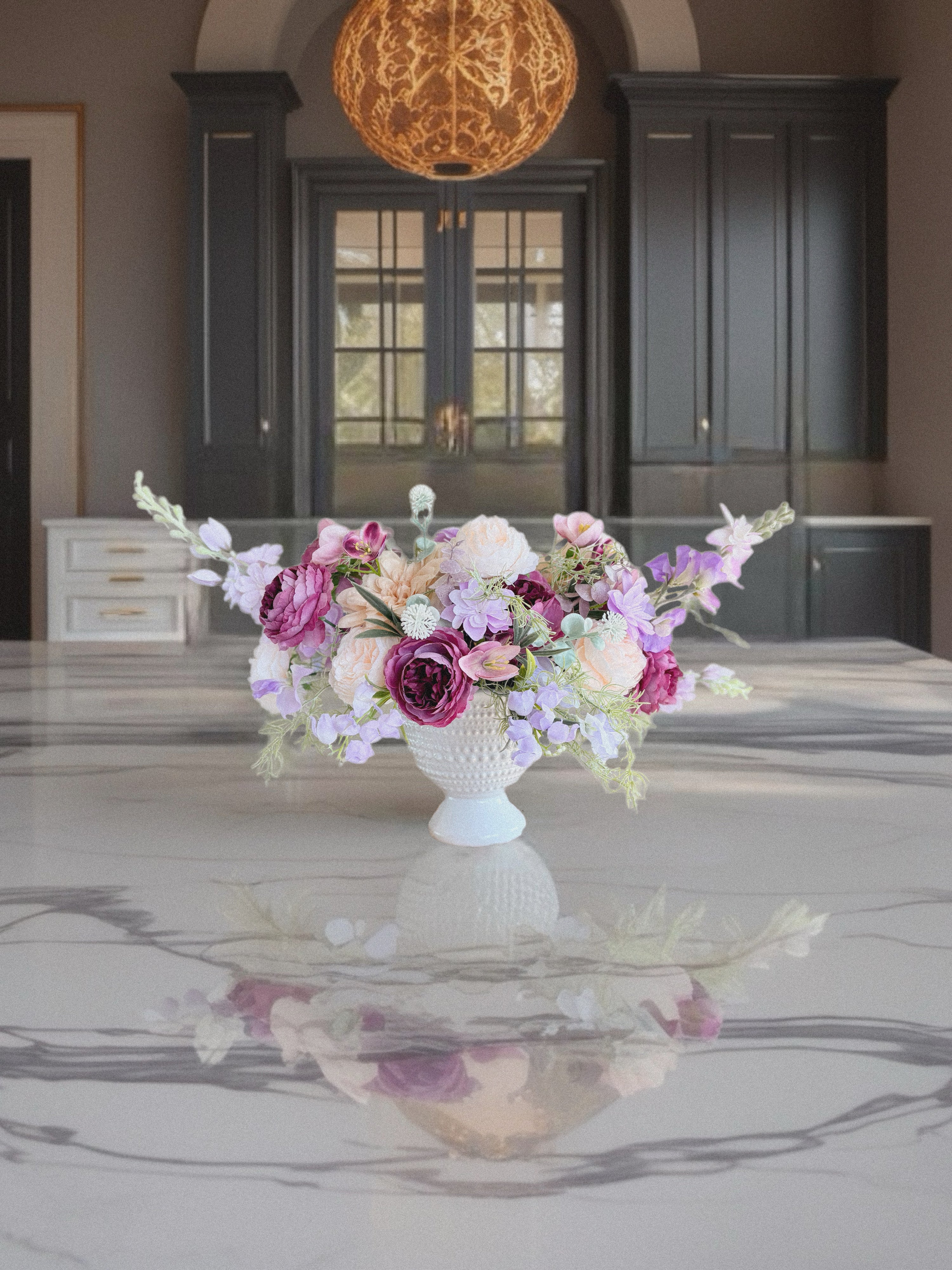 Floral arrangement in a white vase on a reflective surface with dark cabinets in the background