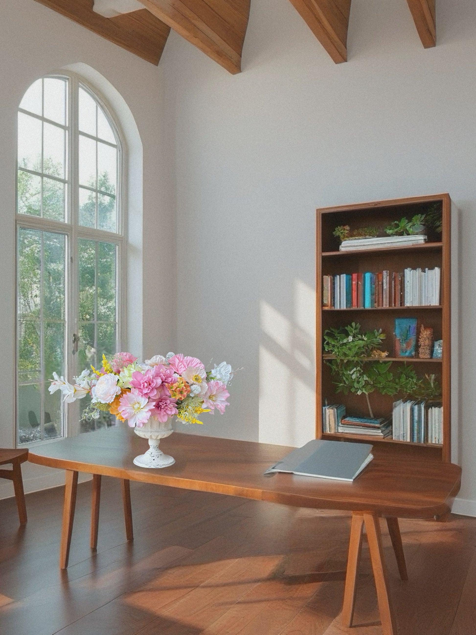 Wooden desk with flowers and a bookshelf in a bright room with large windows.