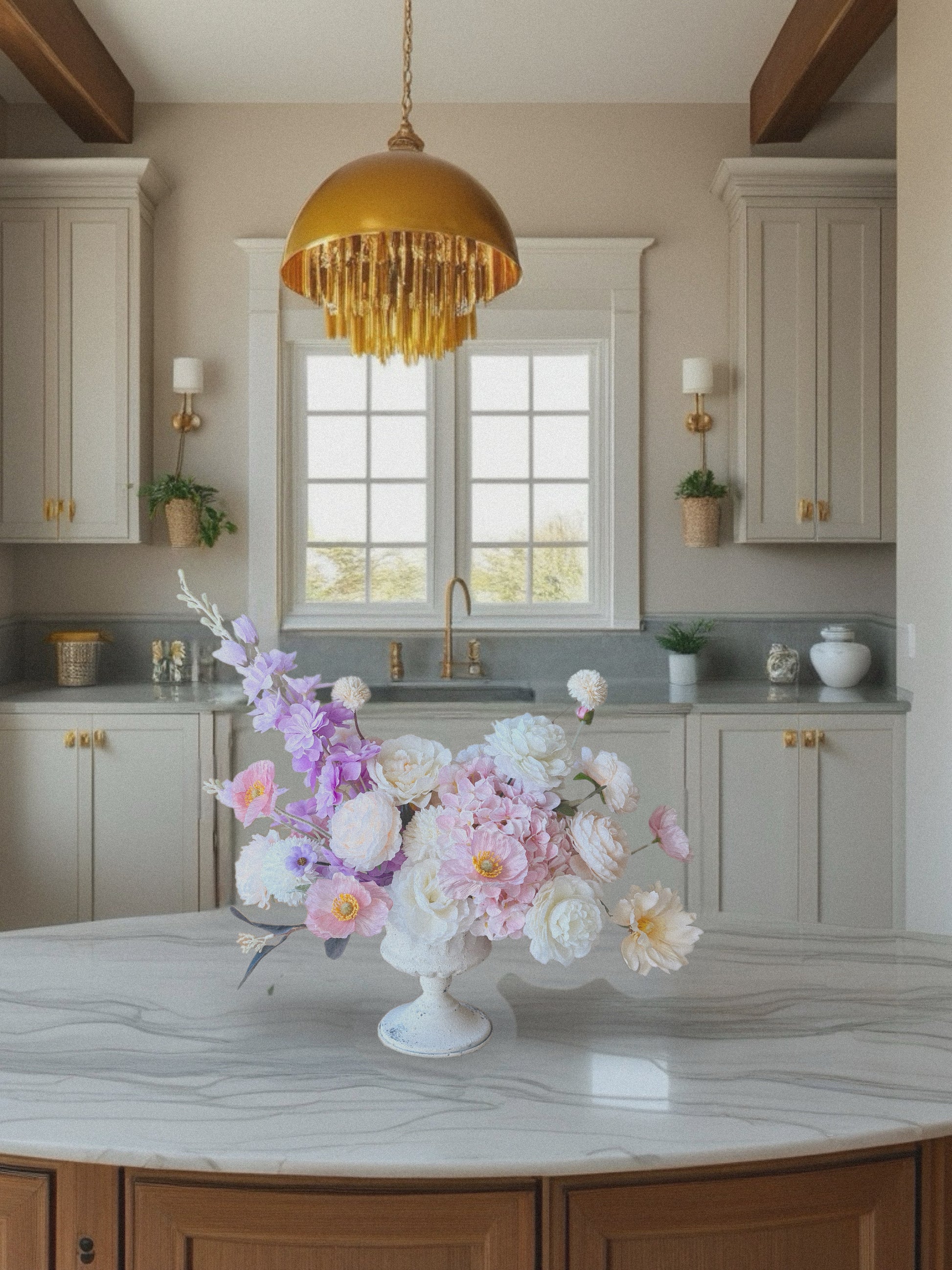 Kitchen with a floral arrangement on a marble countertop, gold pendant light, and white cabinets.