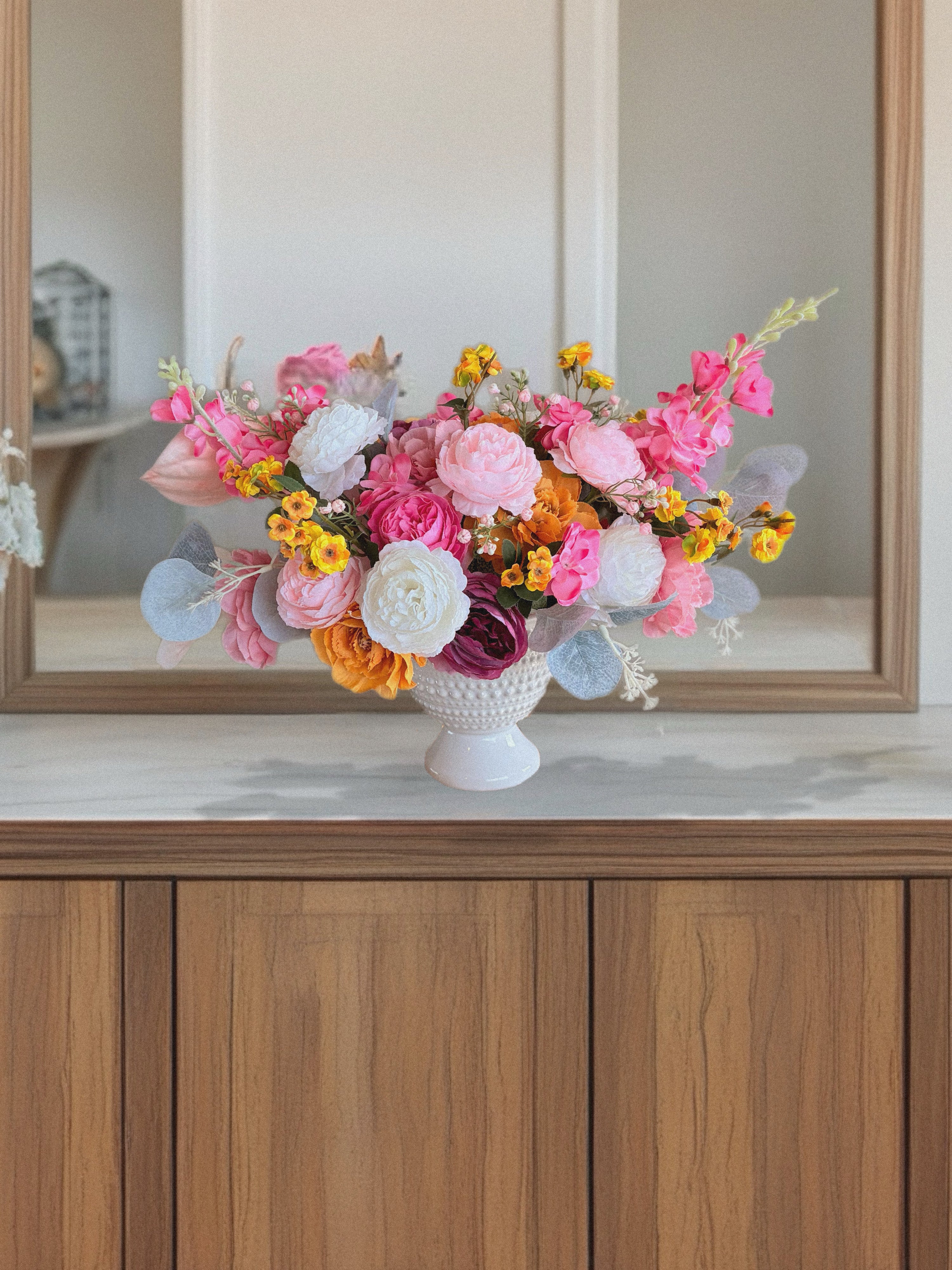 Colorful flower arrangement in a white vase on a wooden surface with a mirror in the background.