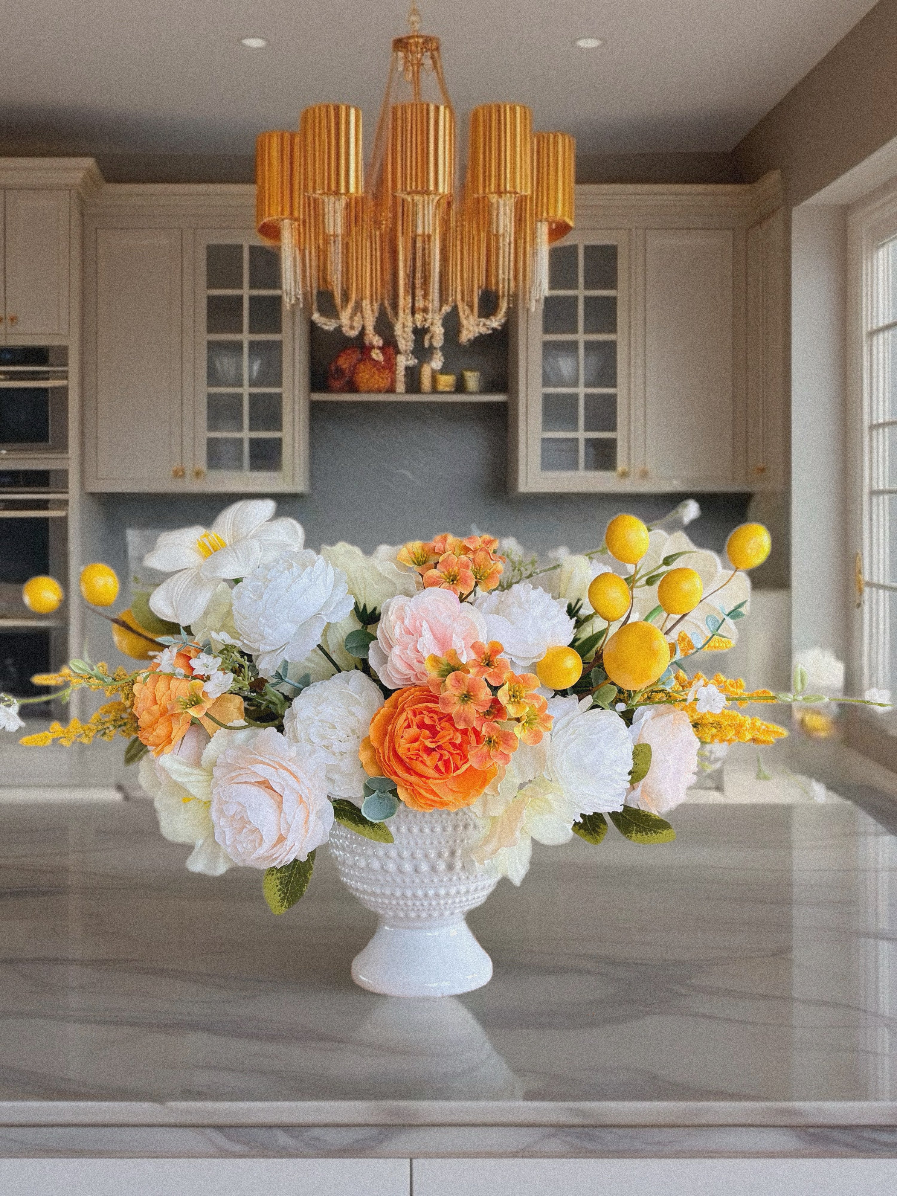 Floral arrangement in a white vase on a kitchen counter with a chandelier above.