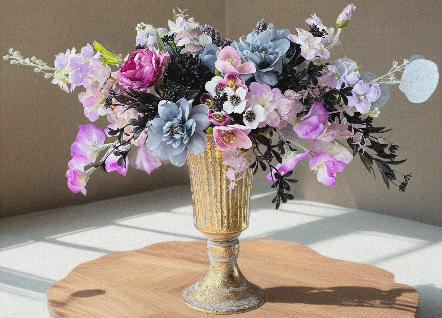 Floral arrangement in a gold vase on a wooden surface with a neutral background