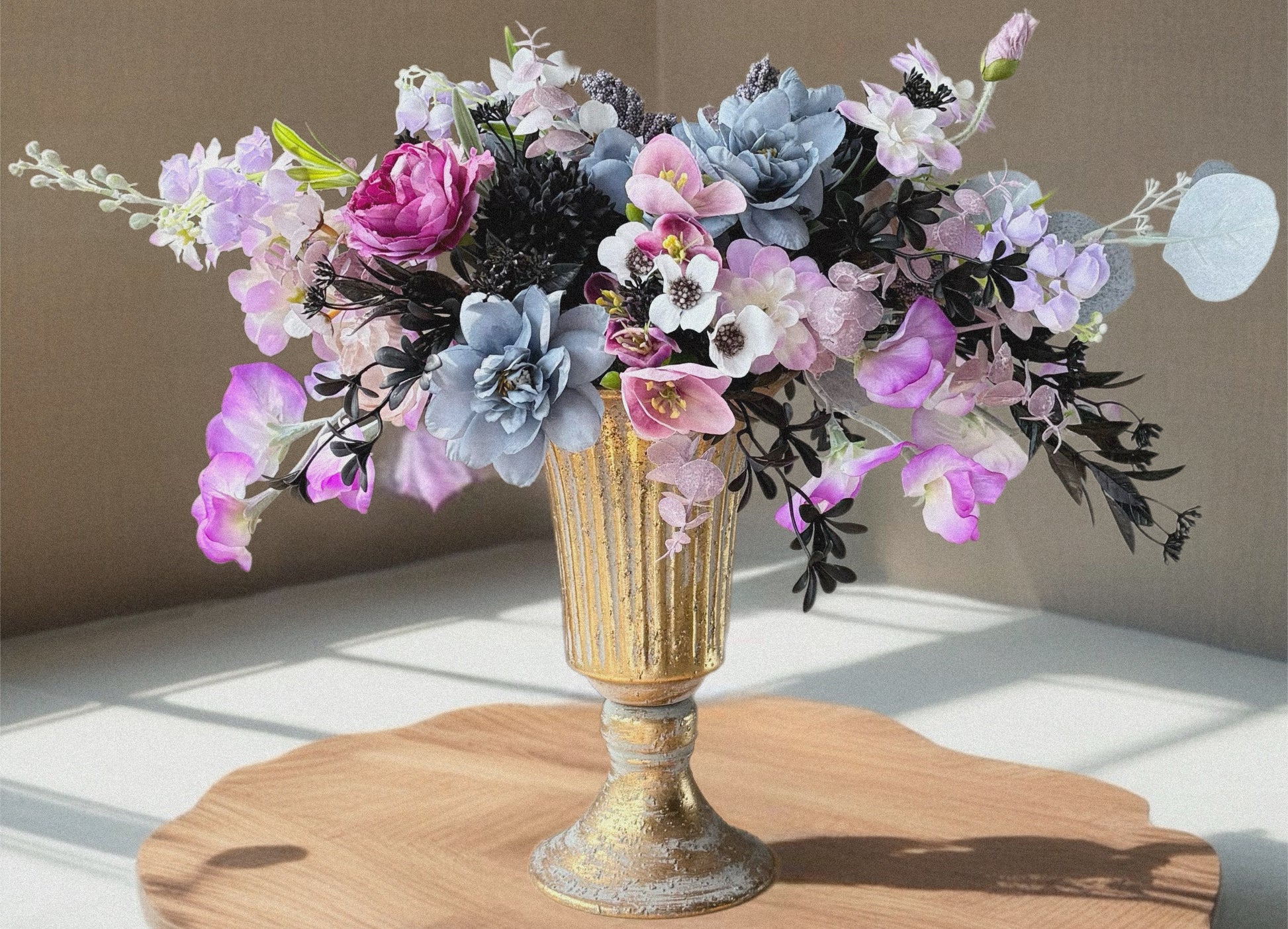 Floral arrangement in a gold vase on a wooden surface with a neutral background
