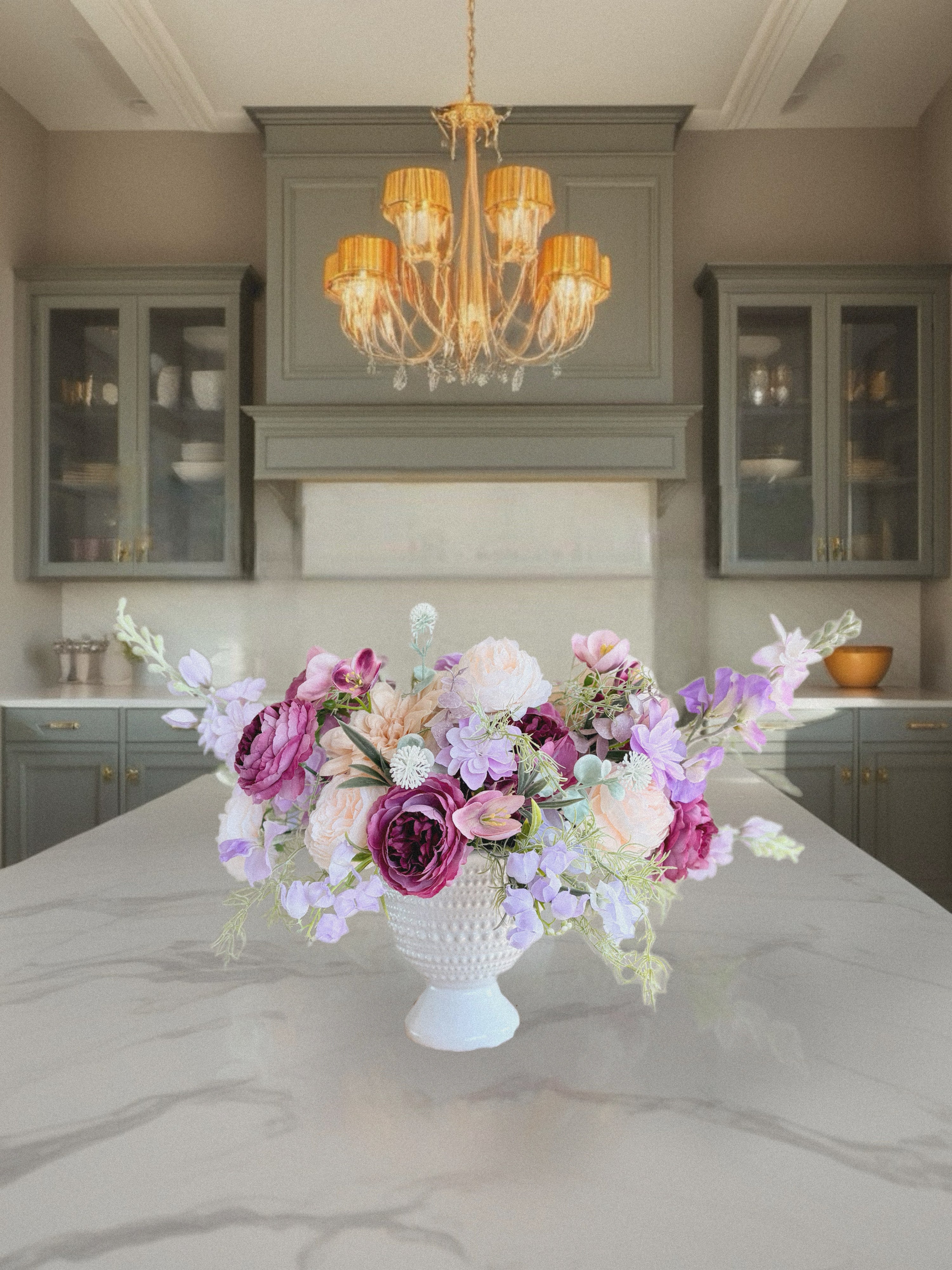 Floral arrangement in a white vase on a kitchen island with a chandelier above.