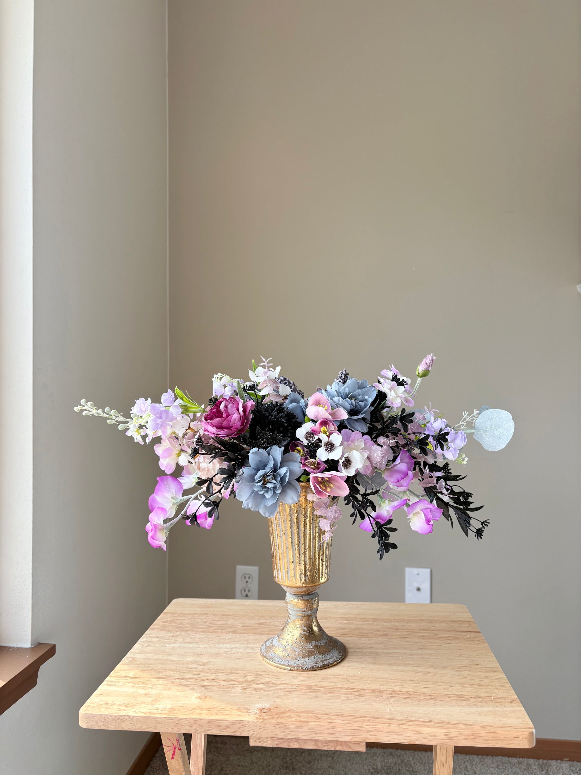 Floral arrangement in a gold vase on a wooden table against a plain wall.