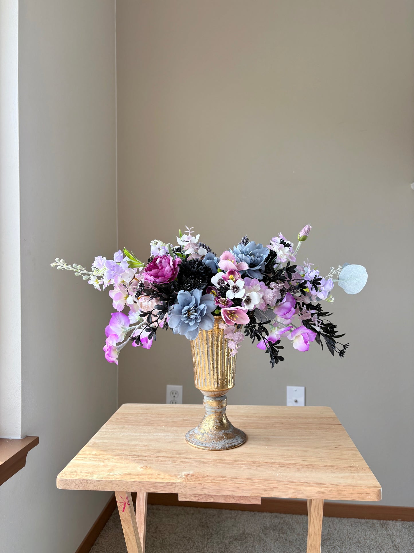 Floral arrangement in a gold vase on a wooden table against a plain wall.
