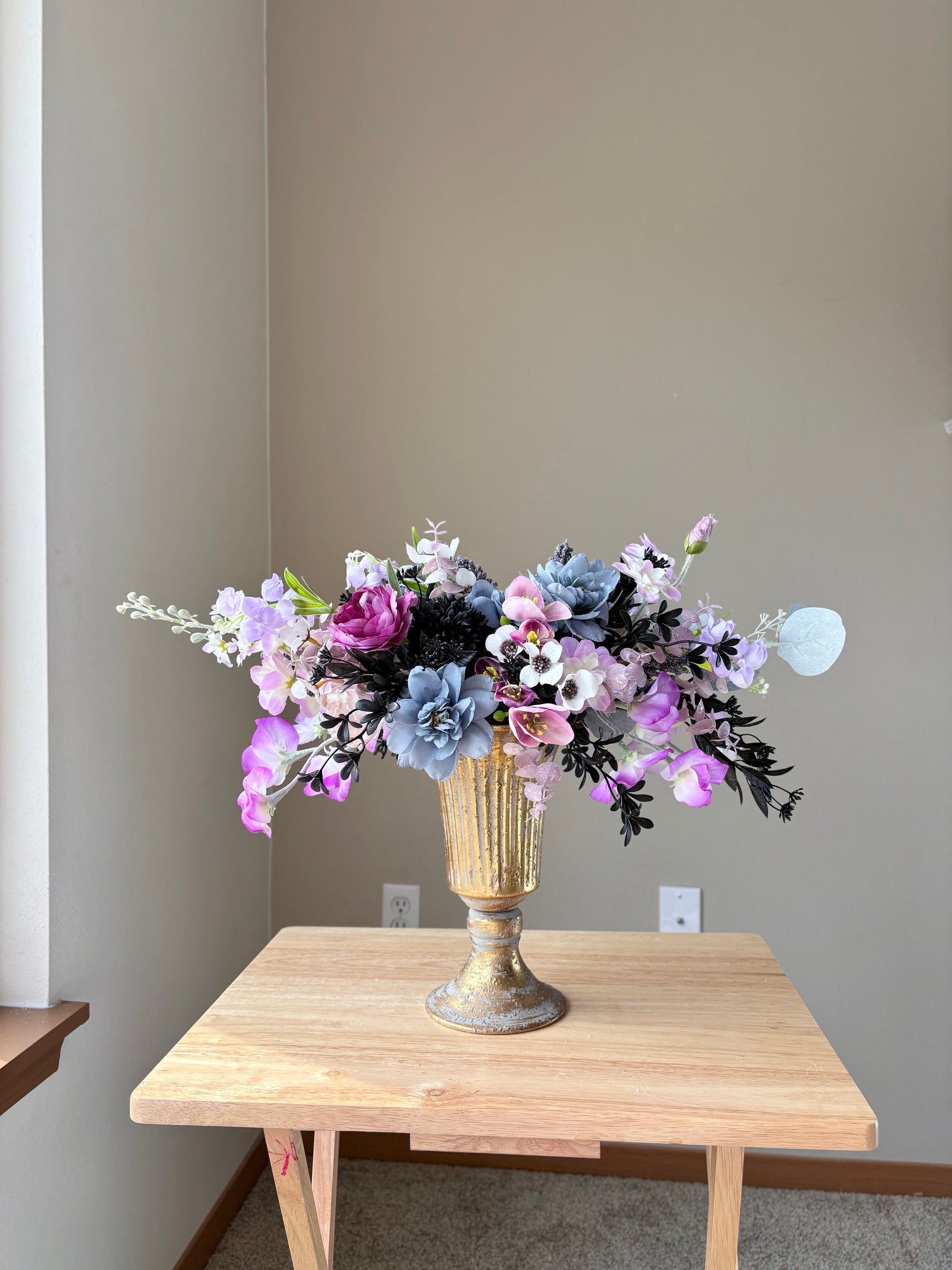 Floral arrangement in a gold vase on a wooden table against a plain wall.