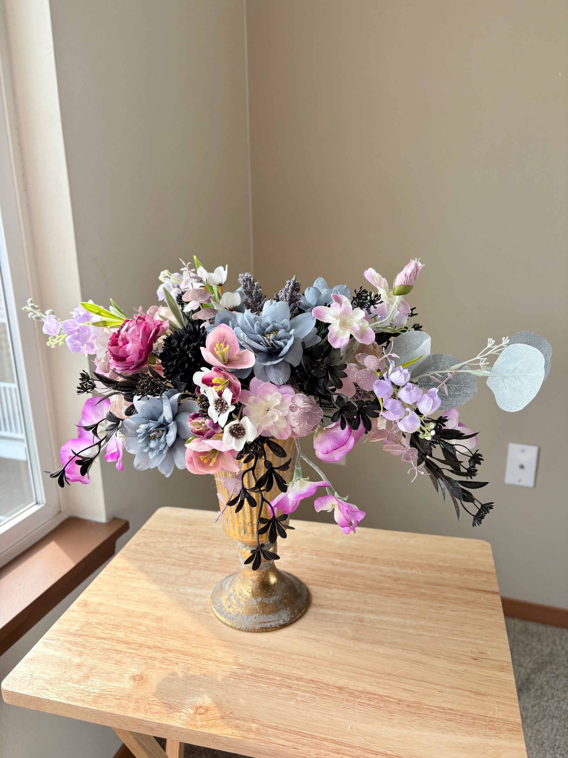 Floral arrangement in a decorative vase on a wooden table near a window.