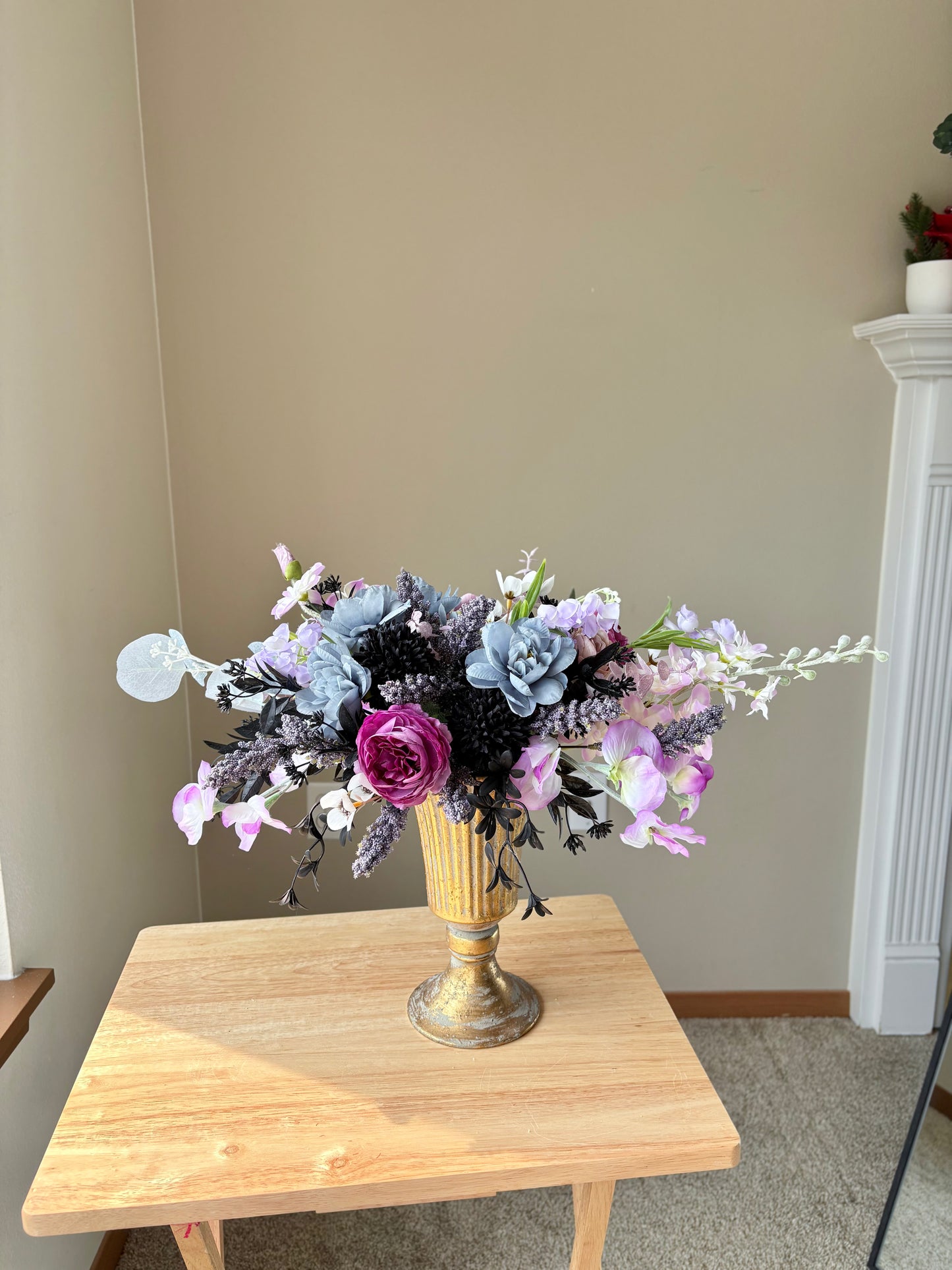 Floral arrangement in a gold vase on a wooden table against a beige wall.