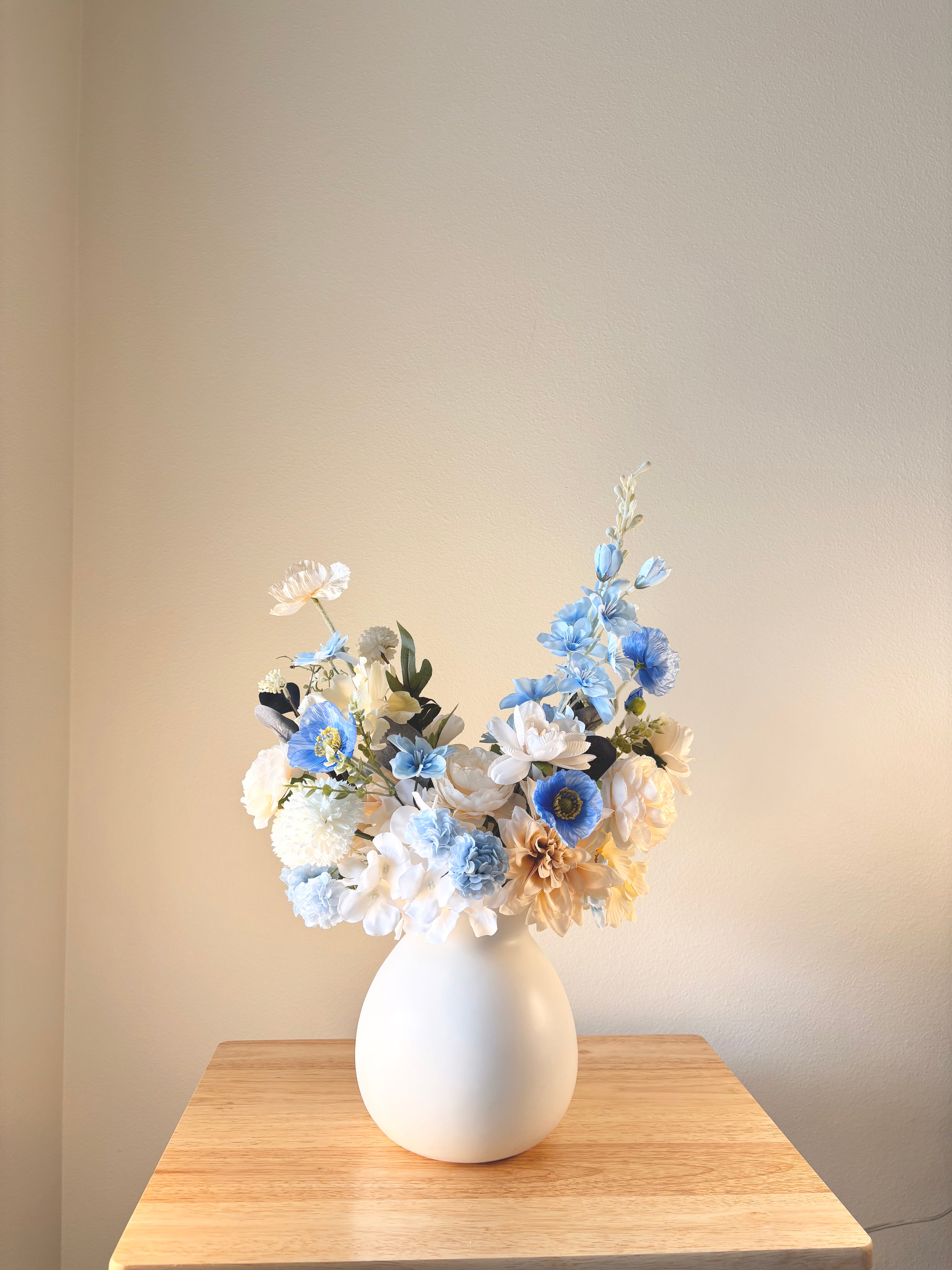 White vase with a bouquet of flowers on a wooden table against a plain wall