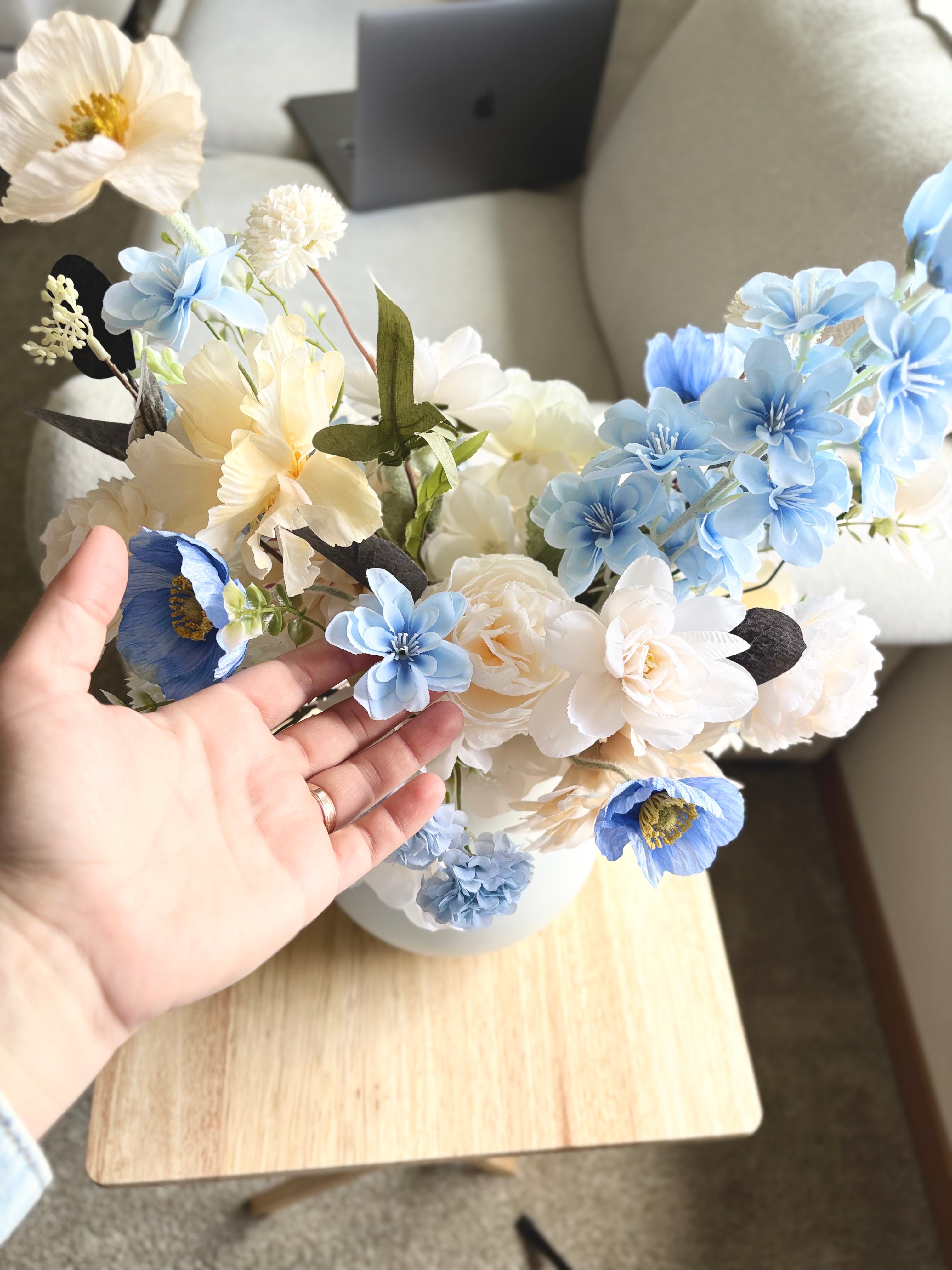 Hand holding a small bouquet of flowers next to a larger arrangement on a wooden surface.