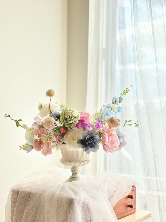 Floral arrangement in a decorative urn on a table with sheer curtains in the background