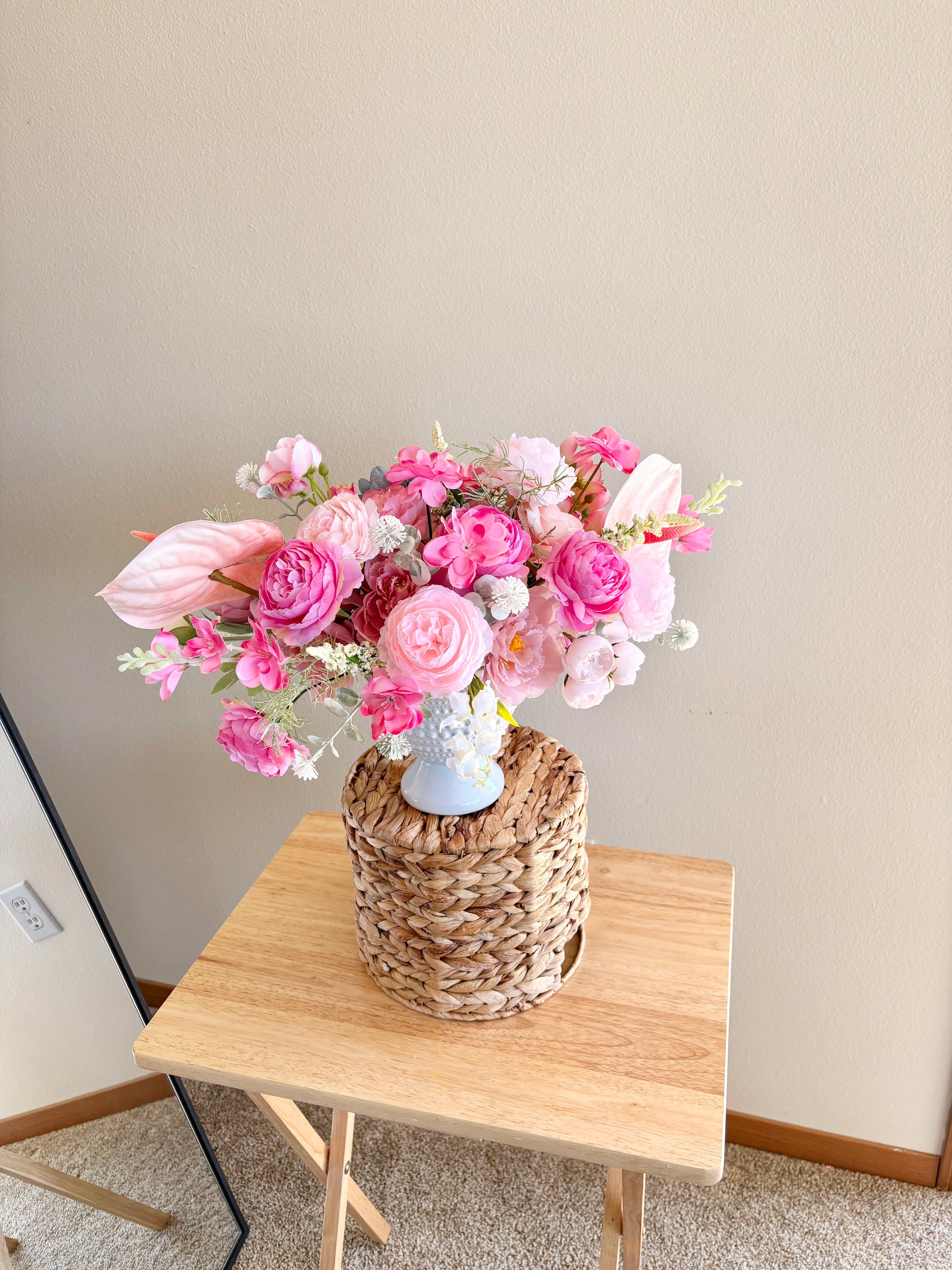 Bouquet of pink flowers in a woven basket on a wooden table against a plain wall.