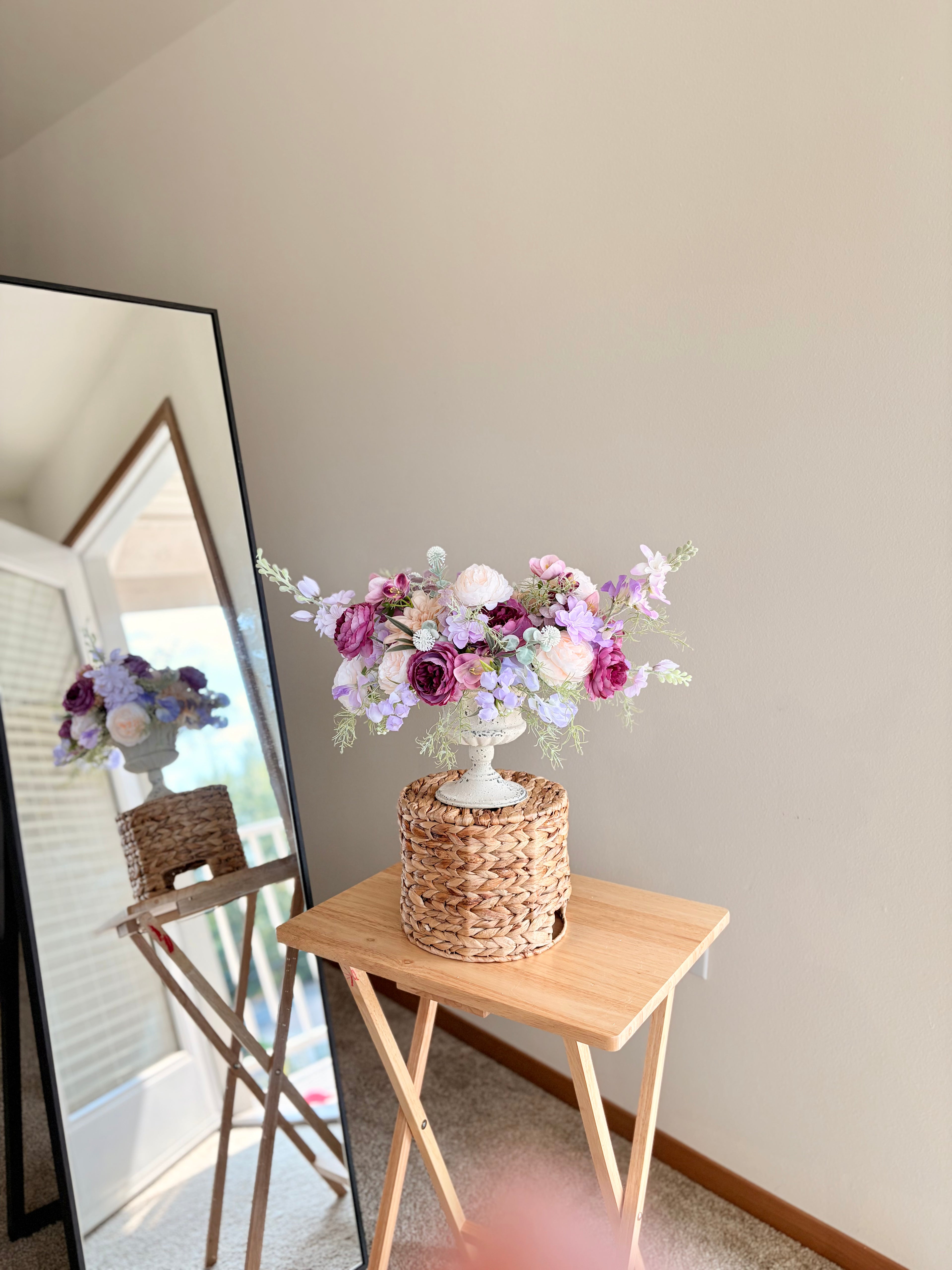 Floral arrangement on a small wooden table with a mirror reflecting a chair and window.