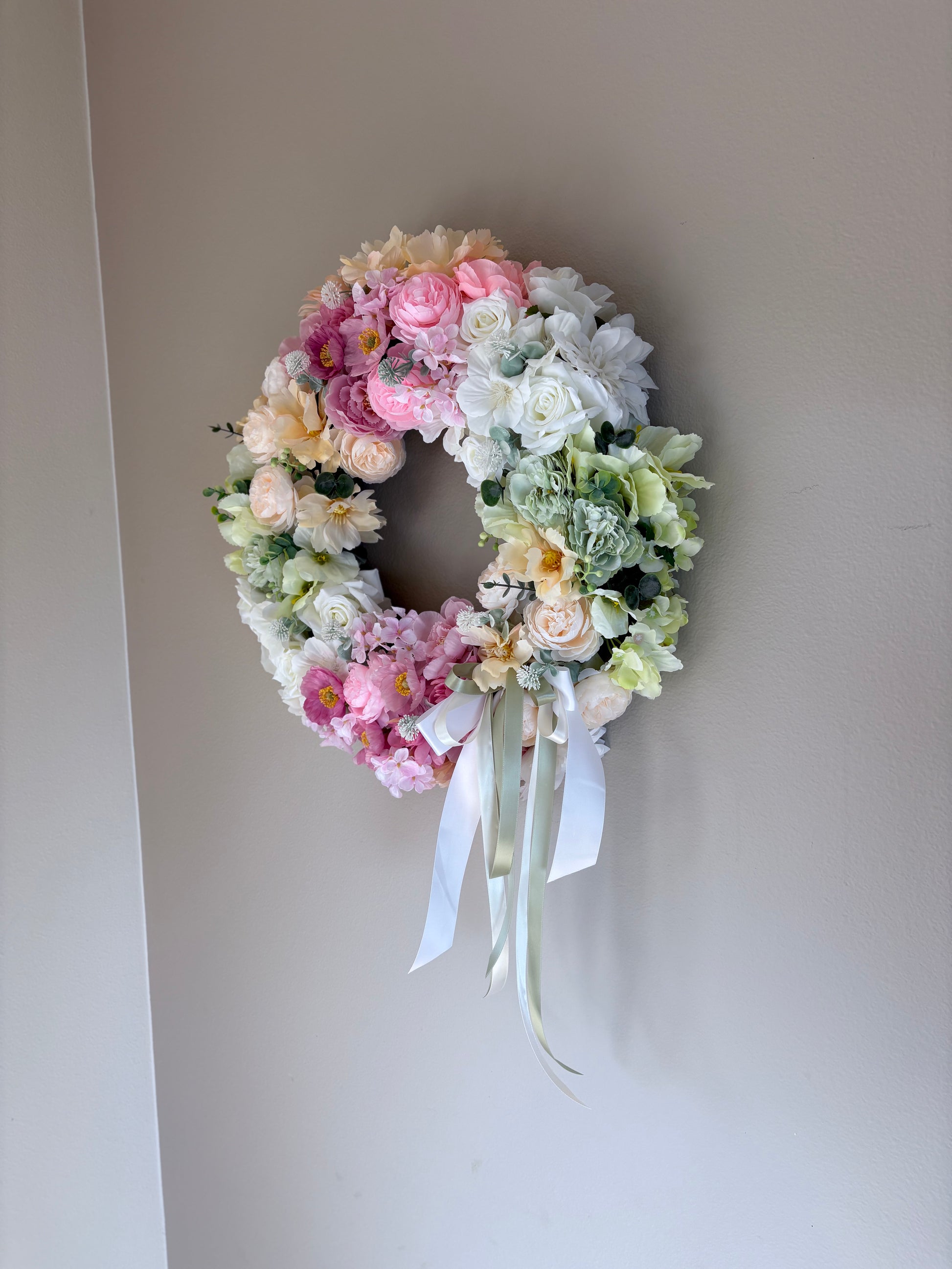 Floral wreath with pink, white, and green flowers on a plain wall.